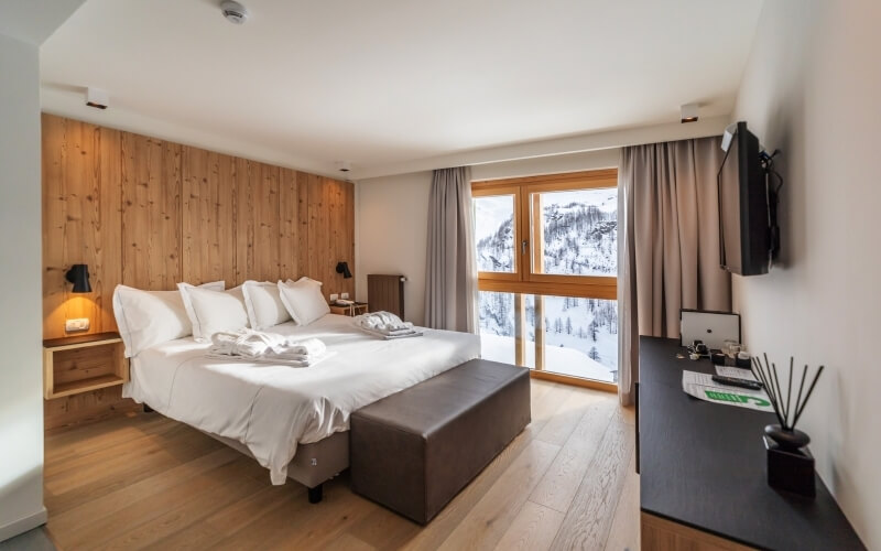 Camera d'hotel moderna con letto grande, scrivania nera e vista panoramica su montagne innevate attraverso una grande finestra.