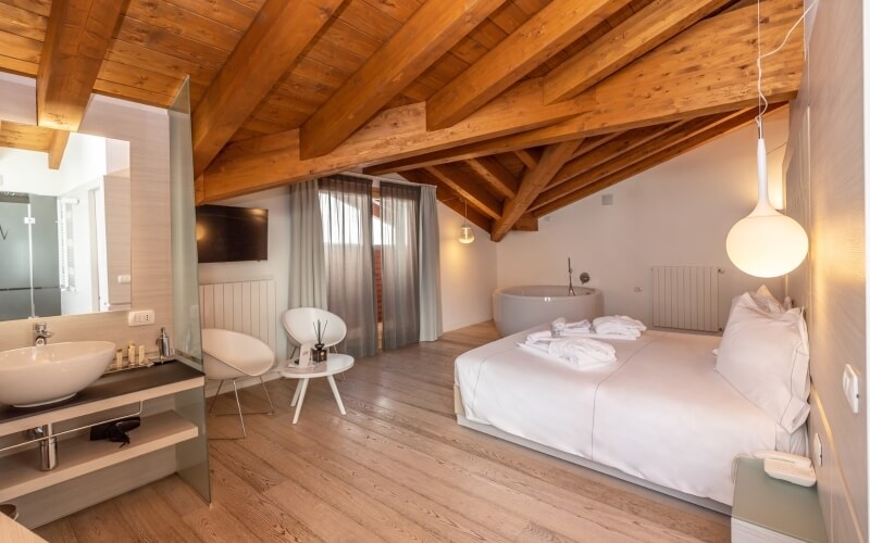 Modern minimalist hotel room with a lofted ceiling, large bed, bathtub, sink, and light wood flooring, exuding calmness.