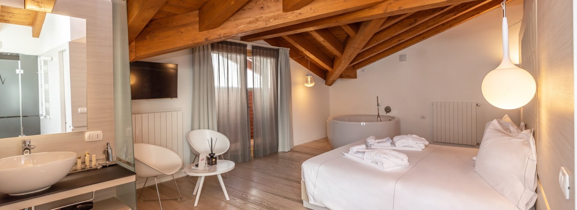 Modern hotel room with sloping ceiling, bathtub, single bed, and minimalist decor, featuring wooden beams and large window.