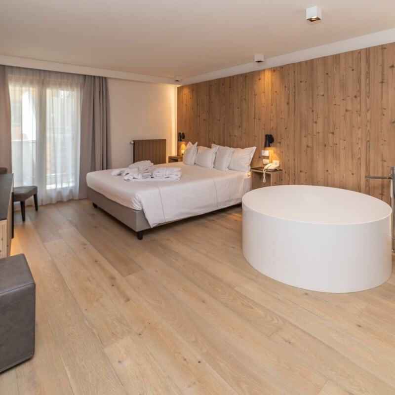 Spacious room with a bed, circular tub, sitting area, and elegant decor, featuring light wood flooring and white walls.