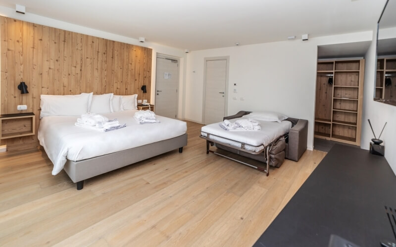 Hotel room featuring a white bed, gray couch, folded laundry, light wood floor, and white walls with shelving.