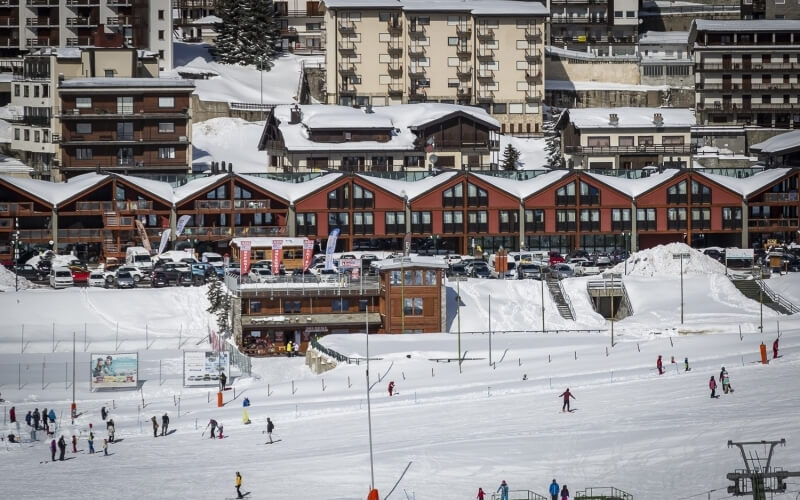 A snowy ski village with skiers, ski lifts, and colorful multi-level buildings nestled on a slope under a clear sky.