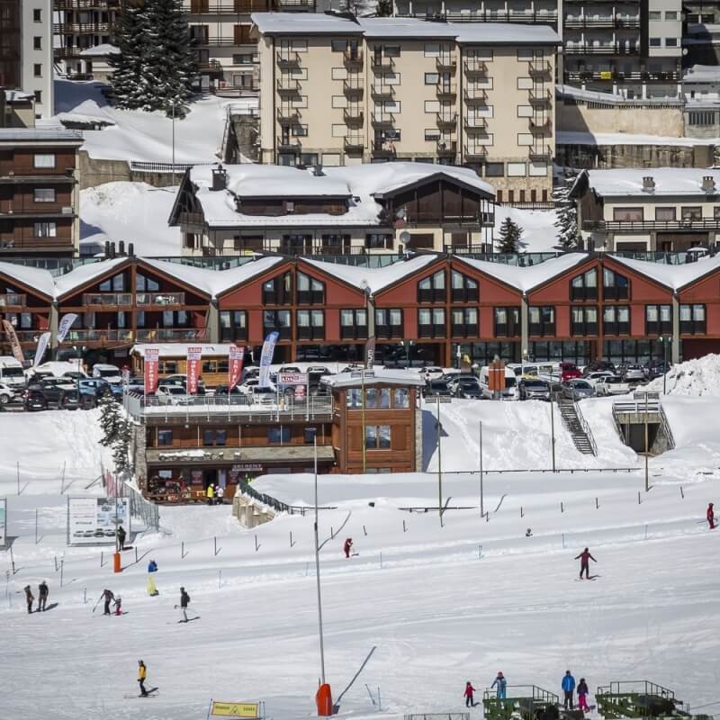 Snowy ski resort town with a red central building, ski slope, skiers, and evergreen trees in a bustling winter atmosphere.