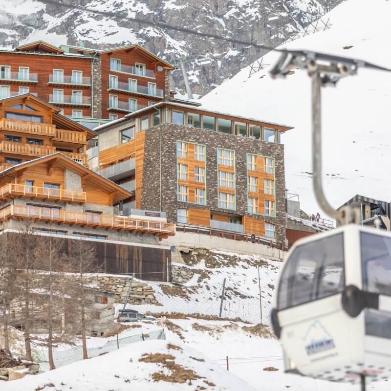 Winter scene of a ski resort with a large hotel, smaller buildings, and a ski lift against snowy mountains.