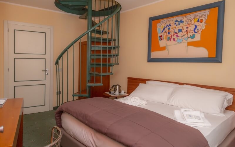 Cozy hotel room with a spiral staircase, double bed, abstract painting, and warm peach walls, inviting relaxation.