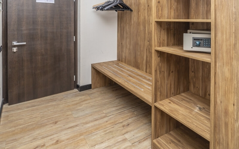 A light wood bench with slatted top and shelves above in a white room, with a dark wood door on the left.