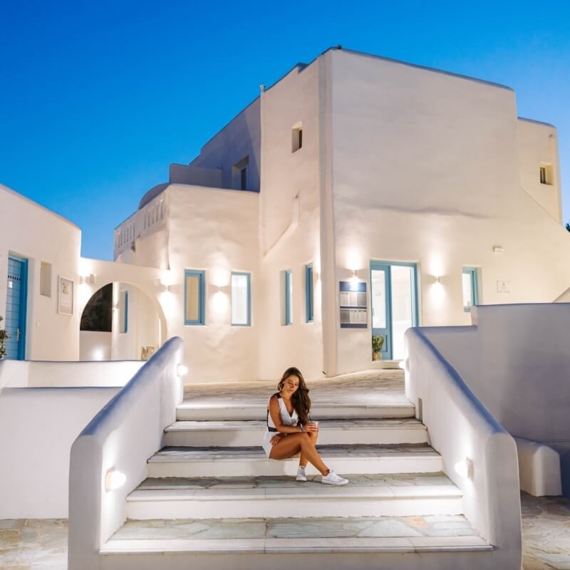 A woman in a white outfit sits on steps of a white building with blue accents, under a clear blue sky, exuding tranquility.