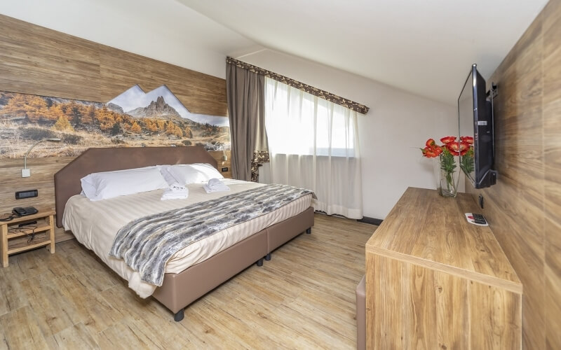 Serene bedroom with a double bed, wooden accents, vibrant flowers, and a mountain view through a large window.