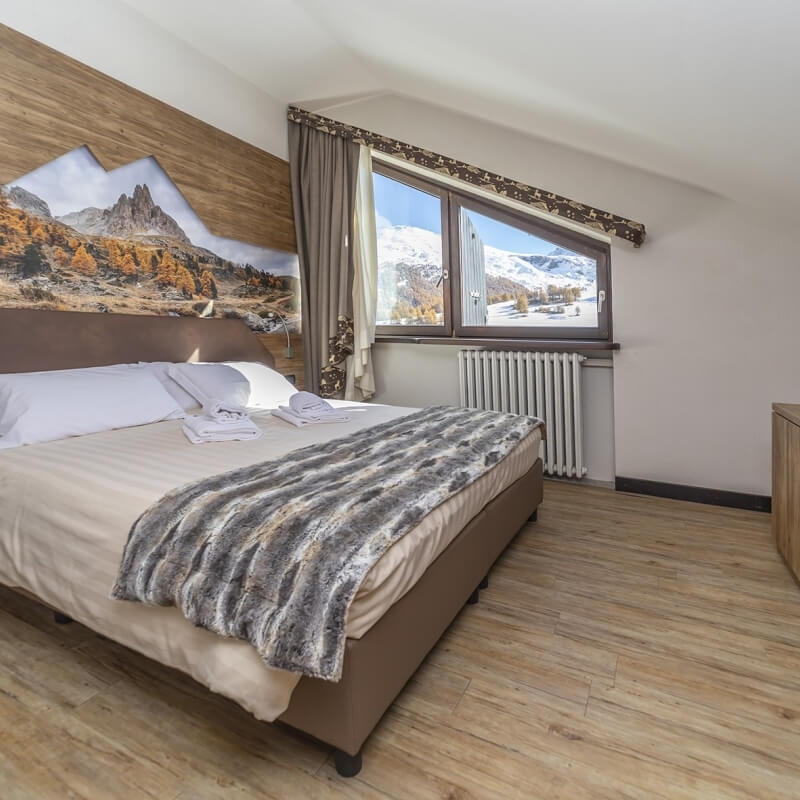 Serene bedroom with a neatly made bed, mountain mural, and window view of snow-capped mountains under a blue sky.