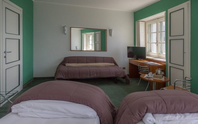 Cozy hotel room with two beds, a couch, desk, and large window, featuring green and white decor and natural light.