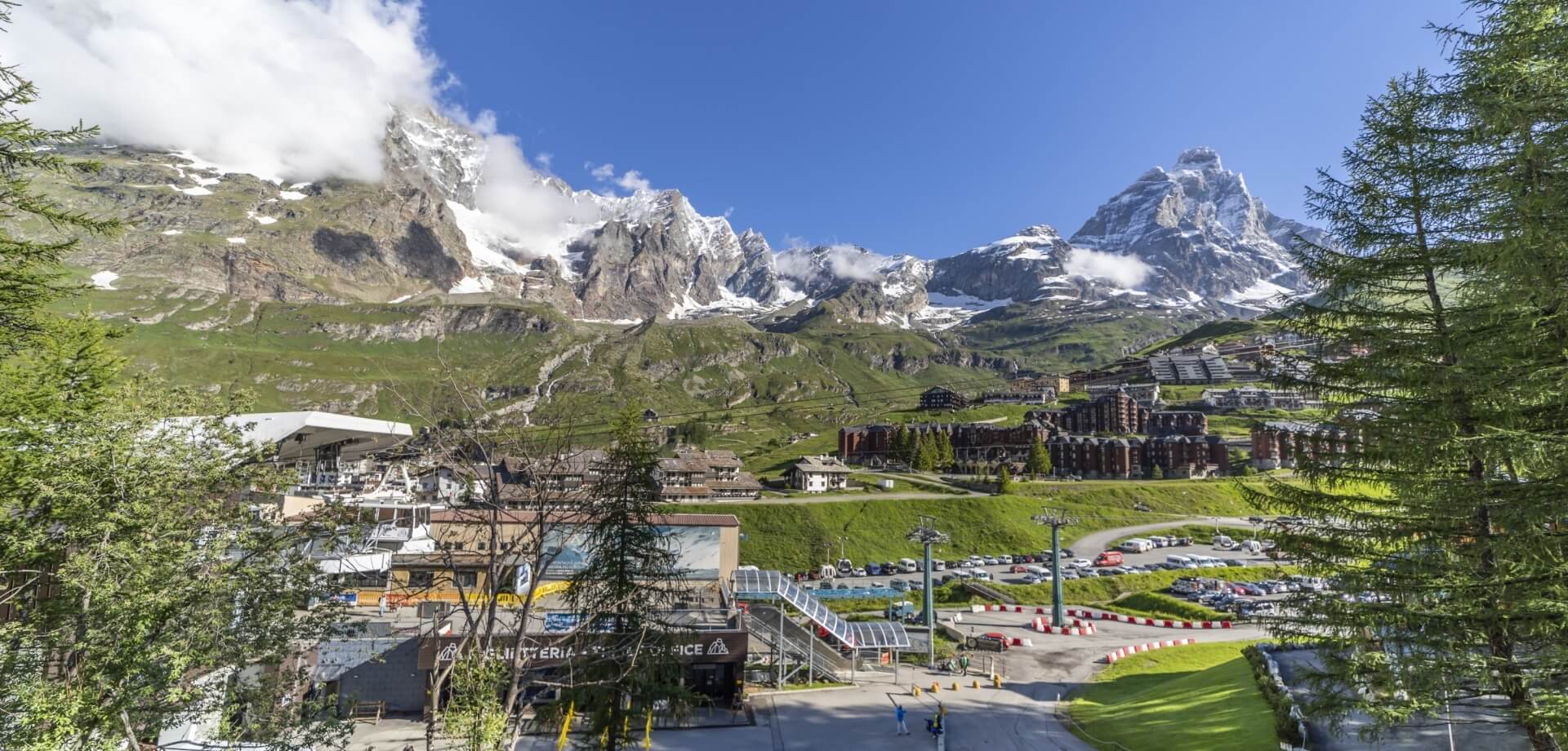 Panorama di un paesaggio montano con un resort sciistico, cime innevate e un cielo blu punteggiato di nuvole bianche.