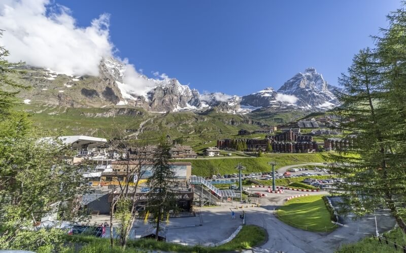 Mountainous landscape featuring a large resort complex, parking lot, trees, and people, suggesting a popular tourist destination.