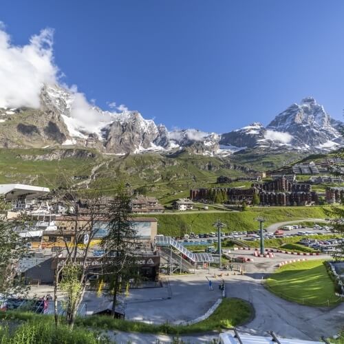 Scenic mountain range with snow-capped peaks, busy tourist area, grassy spaces, and amenities under a clear blue sky.