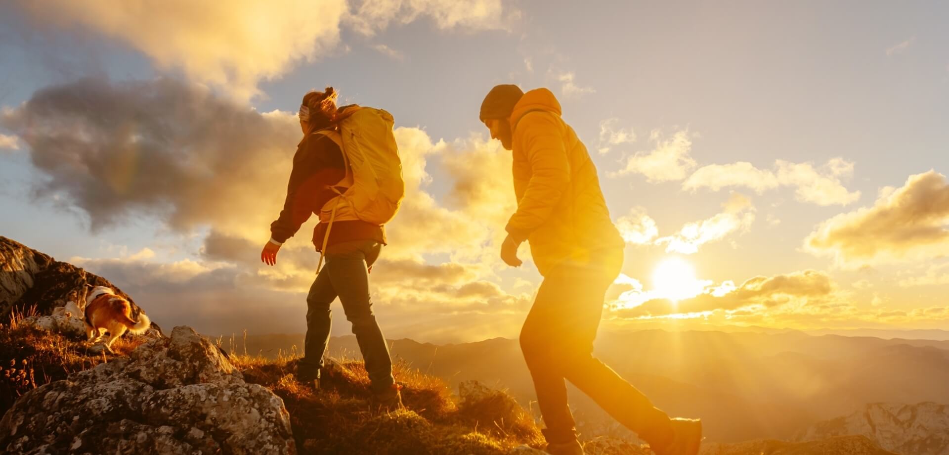 Due persone e un cane su un terreno roccioso, ammirano un tramonto sereno su montagne e nuvole.
