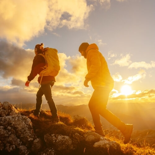 Two individuals climb a grassy hill at sunset, with mountains in the background; one wears a backpack.