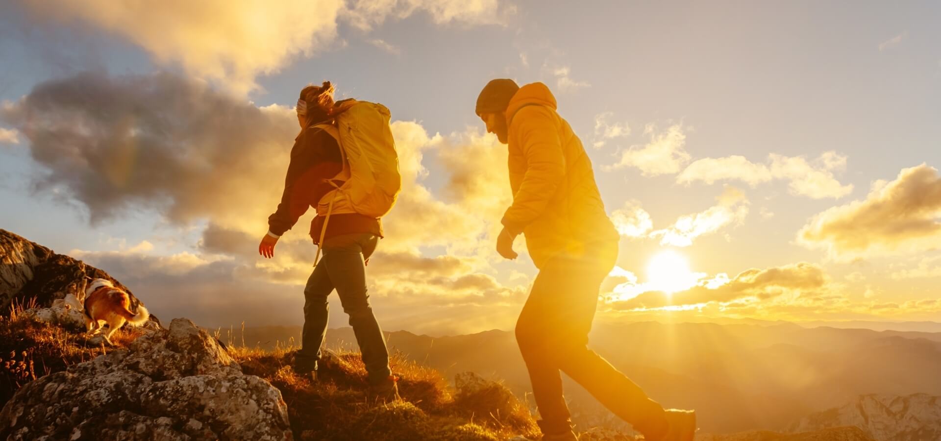 Two hikers and a dog on a mountain summit at sunset, with vibrant clouds and a warm atmosphere.