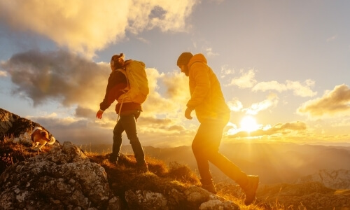 Two hikers in warm clothing ascend rugged terrain, with a small dog in the background and a setting sun behind them.