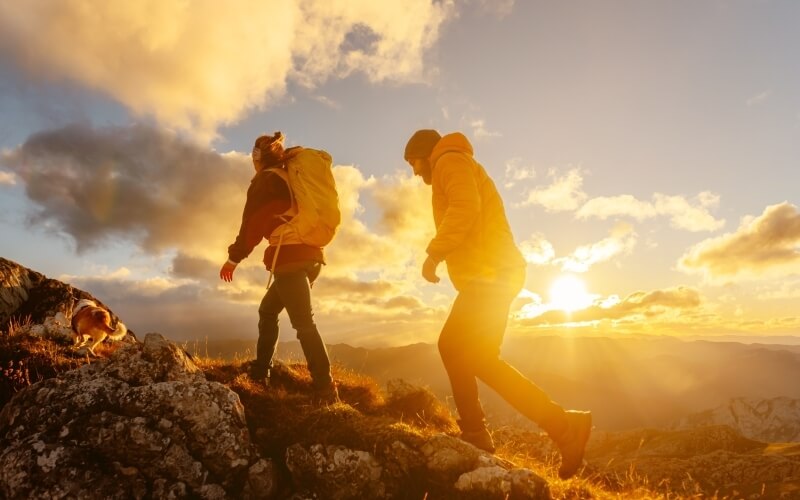 Due escursionisti con un cane bianco e macchie marroni camminano su una montagna al tramonto, circondati da natura serena.