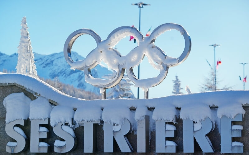 Winter scene featuring snow-covered Olympic rings and "SESTRIERE" text, with trees and streetlights under a clear blue sky.