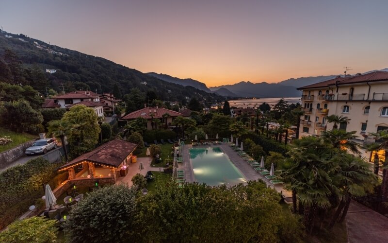 Serene resort scene with a pool, red-roofed buildings, trees, mountains, and a distant car under a clear sky.