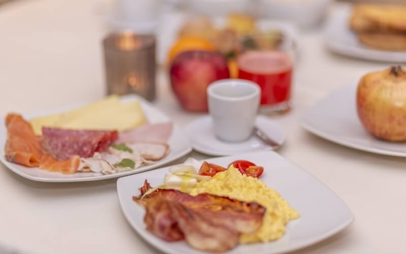 Elegant hotel breakfast setting with scrambled eggs, bacon, meats, cheeses, fruit, juice, and a lit candle on the table.