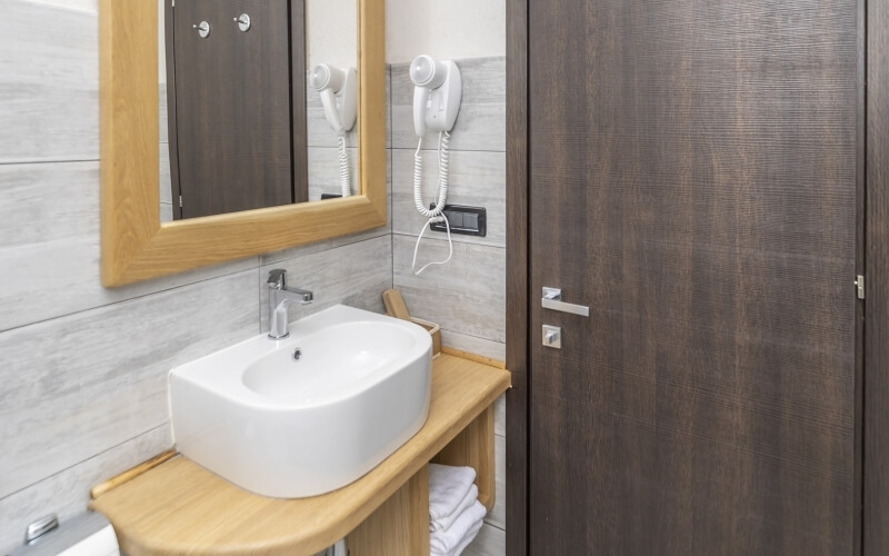 Bagno moderno con lavabo bianco, specchio, asciugacapelli e asciugamani piegati su un ripiano in legno.