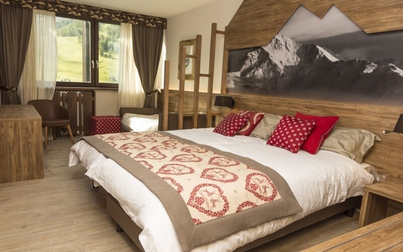 Serene bedroom with mountain-themed decor, large window, double bed with red pillows, and wooden desk overlooking nature.