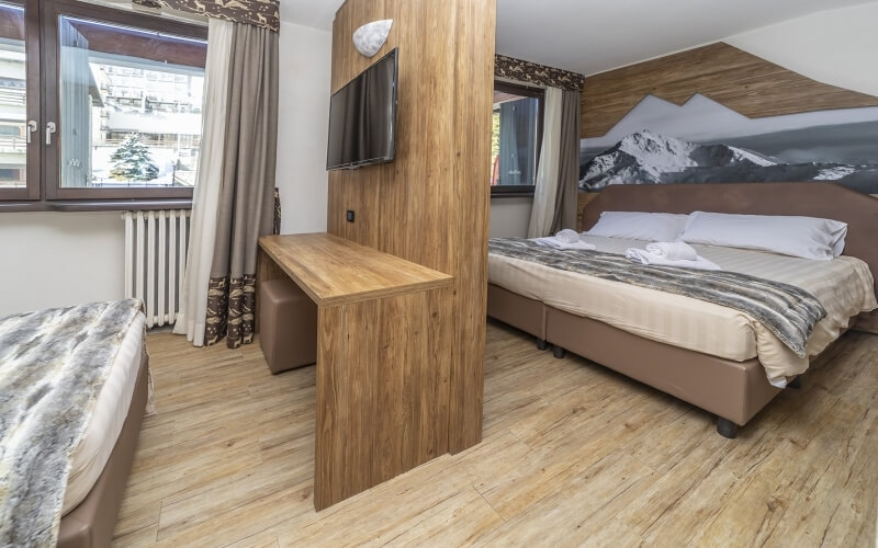 Modern hotel room with two beds, a desk, light wood floors, mountain painting, and a cozy atmosphere.