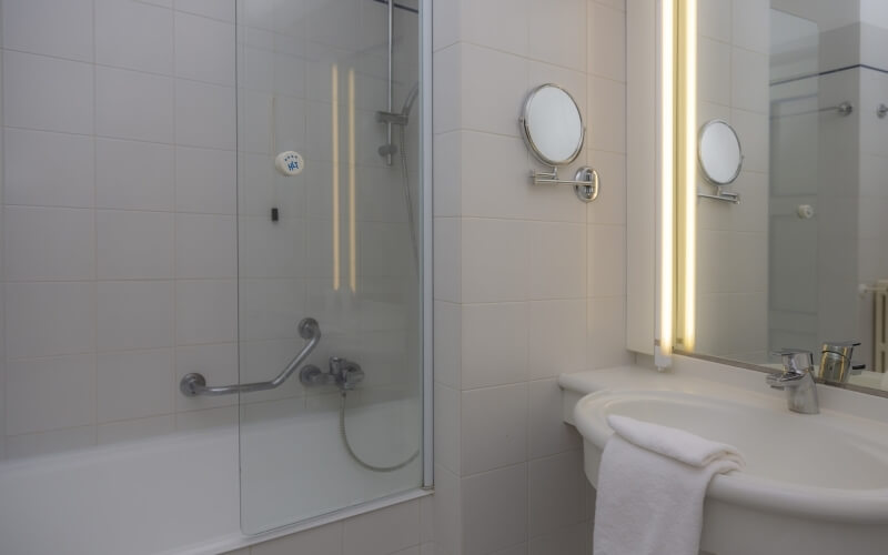 Bagno moderno con doccia in vetro, vasca, lavandino, asciugamano bianco e specchi rotondi su pareti bianche.