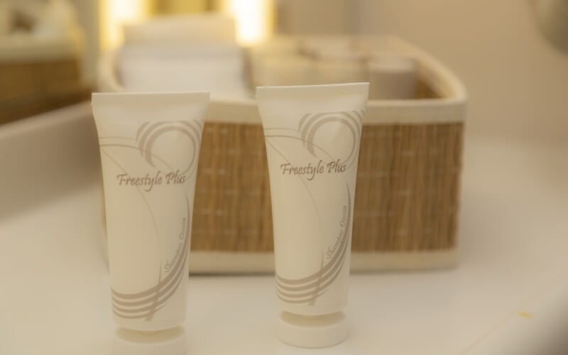 Two white tubes of Freestyle Plus toothpaste on a white surface, with a blurred tan basket in a softly lit, cozy setting.