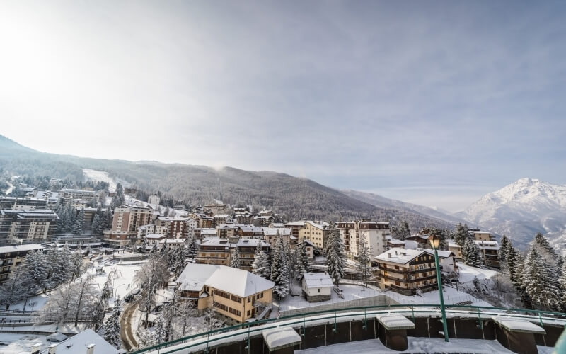 Una città innevata ai piedi delle montagne, con edifici, alberi e un ponte che collega diverse parti del paese.