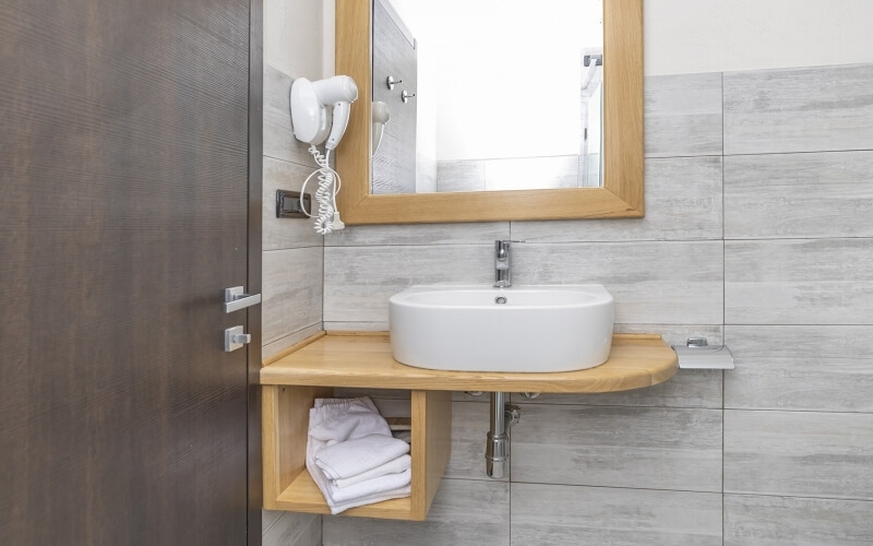 Bathroom scene with a light wood vanity, white sink, mirror, hair dryer, and tiled walls, exuding cleanliness and minimalism.