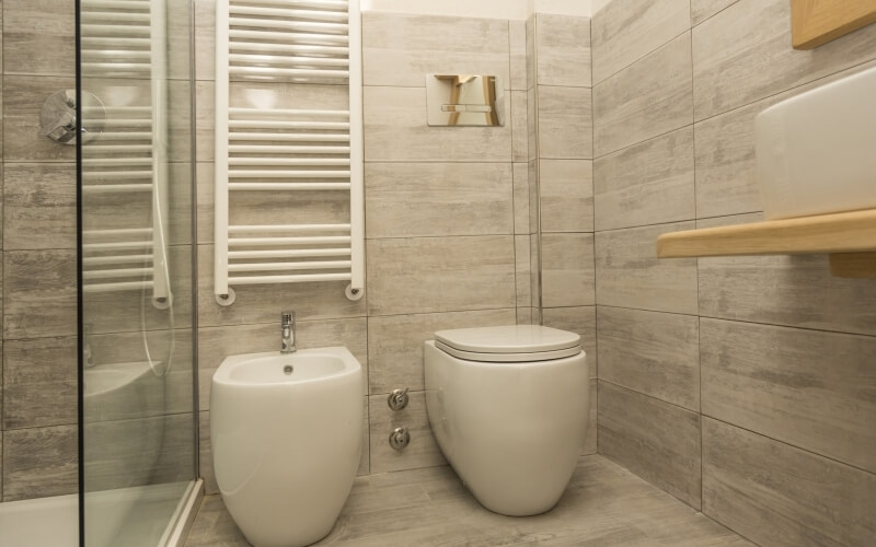 Modern bathroom with a toilet, bidet, shower, light gray tiled walls, large floor tiles, and a wooden shelf above the toilet.