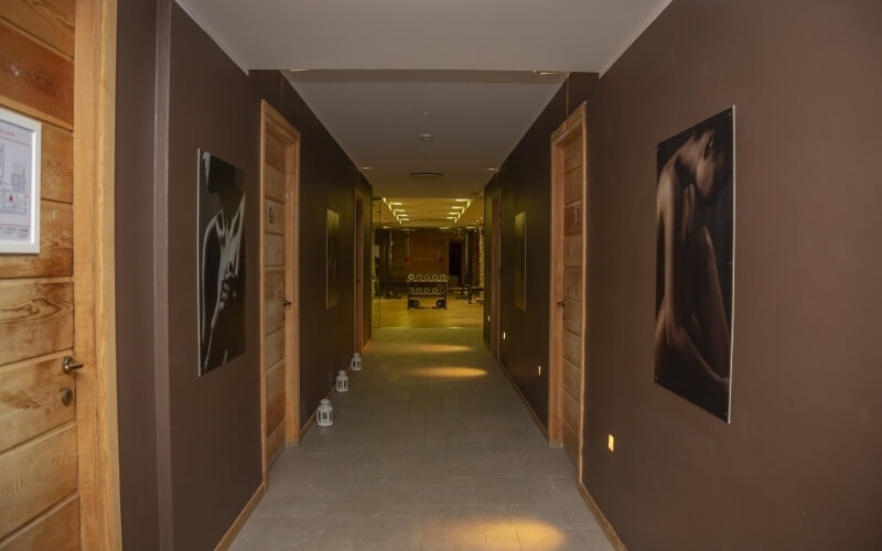 Long hallway with brown walls, wooden doors, black-and-white photographs, gray tiled floors, and exercise room in the background.