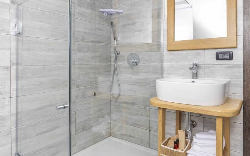 Modern bathroom featuring a glass shower stall, white sink on a wooden table, and light gray tiled walls with dark stripes.