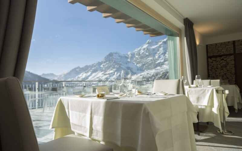 Bright restaurant with white decor and large window showcasing snow-covered mountains, set with elegant tables and chairs.