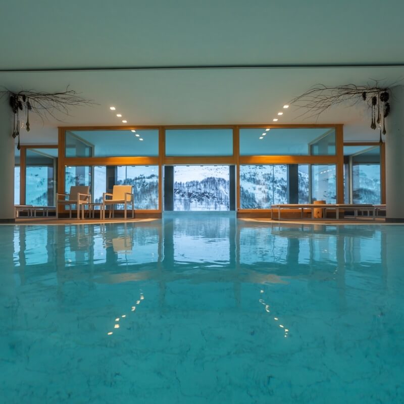 Indoor pool scene with reflective water, wooden-framed windows, snow-covered mountains, and cozy furniture, exuding tranquility.