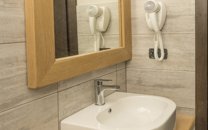 Bathroom sink with a light-wood countertop, large mirror with blow dryers, and dark-brown wooden pillar on the right.