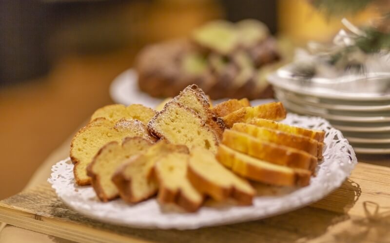 Sliced pound cake on a decorative white plate, arranged on a wooden table with a blurry background of other food items.