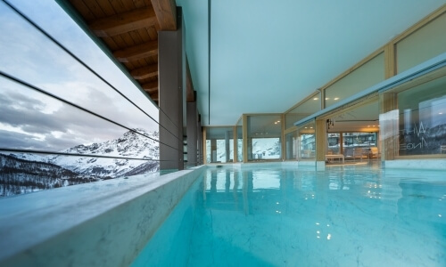 Indoor pool with marble wall, wooden pillars, and mountain view; blue water reflects warm lighting and snowy peaks outside.