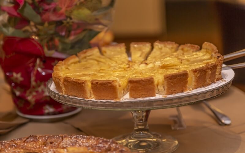 Table with a glass cake stand holding a sliced pie, a vase of flowers, and a glimpse of another dessert.