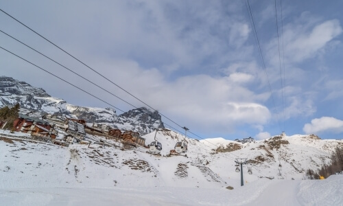 Paesaggio invernale sereno con montagne innevate, un impianto di risalita e edifici sotto un cielo blu con nuvole bianche.