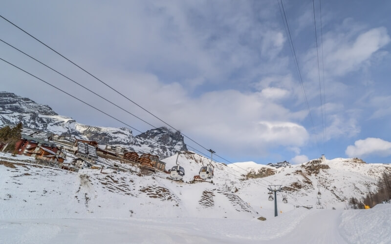 Ski resort at snowy mountain foothills with log cabin-style buildings, empty ski lift, and low clouds casting shadows.