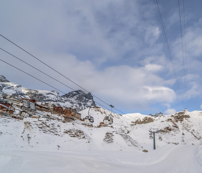Snow-covered mountain range with a ski lift, buildings, power lines, and a blue sky with white clouds.