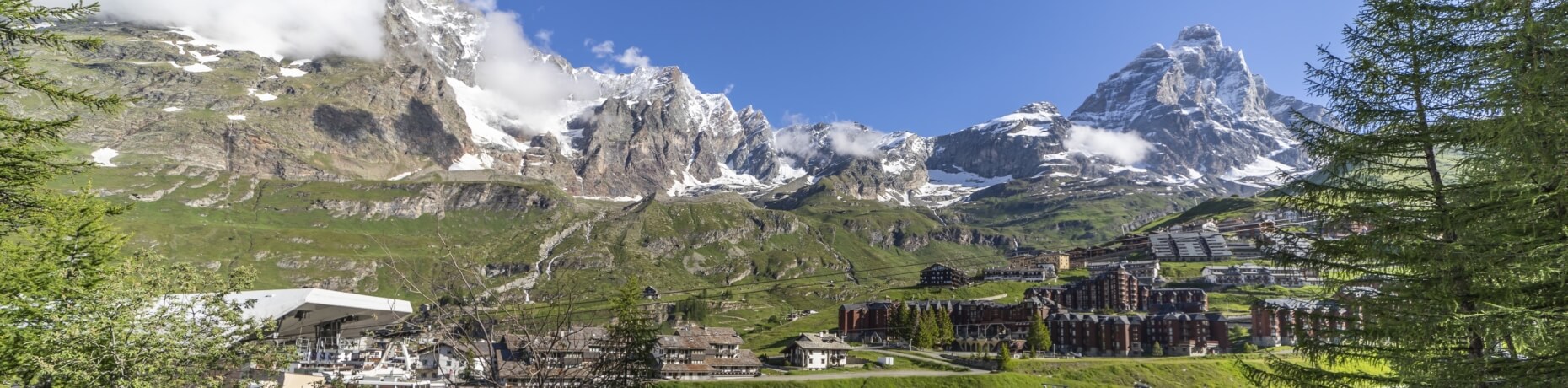 Serene alpine landscape with snow-capped mountains, a charming town, and lush green hills under a brilliant blue sky.