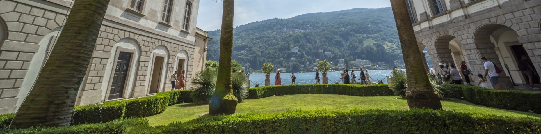 A serene lakeside scene featuring a stone building, lush greenery, people enjoying the view, and a majestic mountain range.