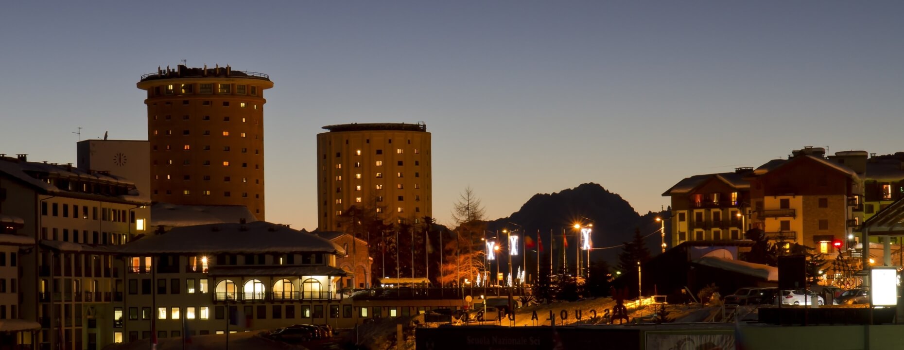 Paesaggio urbano al crepuscolo, edifici illuminati, cielo blu scuro, atmosfera tranquilla con persone e veicoli.