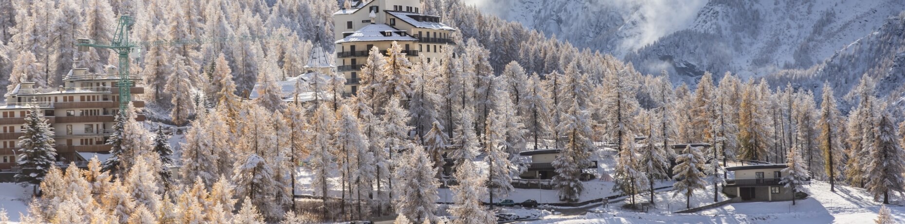 Serene winter scene with multi-story buildings among snow-covered trees and mountains, featuring a crane and parked cars.