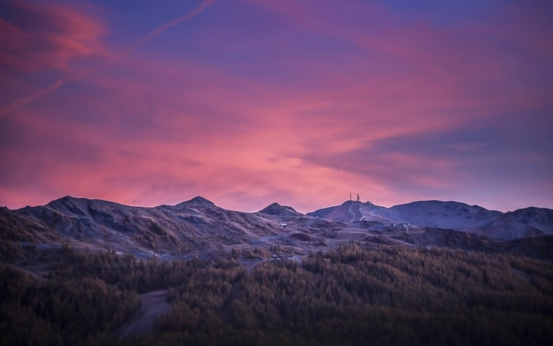 Serene mountain range at sunset, with rugged peaks, scattered trees, and a vibrant pink and purple sky.