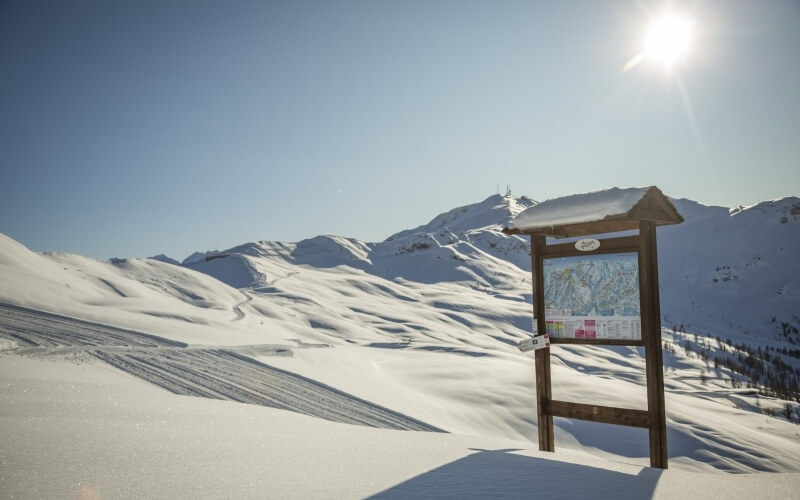 Paesaggio montano innevato con una bacheca in legno e montagne sullo sfondo, cielo blu e sole splendente.